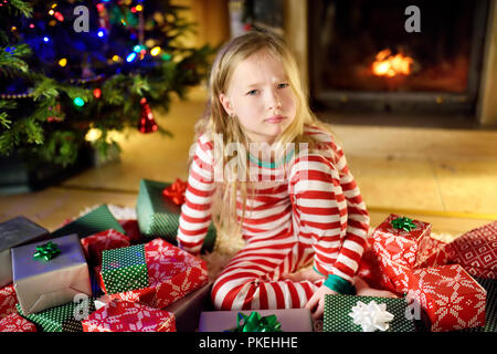 Carino bambina sensazione infelice con i suoi doni di Natale. Bambino seduto da un camino in un accogliente buio in salotto con la vigilia di Natale. Troppi presenta per Foto Stock