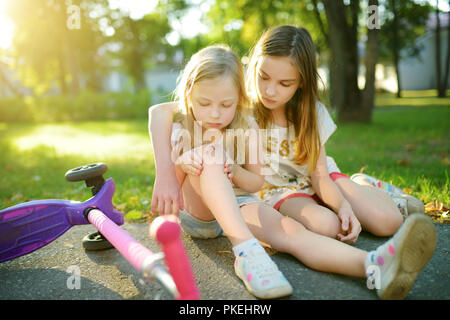 Adorabile ragazza consolante la sua sorellina dopo che lei è caduto il suo scooter a summer park. Bambino di farsi male mentre cavalcate un kick scooter. Famil attivo Foto Stock