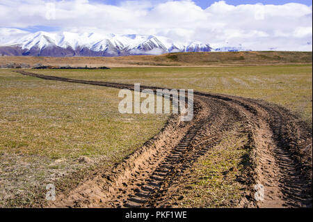 Pneumatico fangoso le piste si snodano attraverso terreni agricoli verso distante mountain range. Alpi del Sud, Nuova Zelanda. Paesaggio rurale, verdi campi e neve picchi placcati Foto Stock