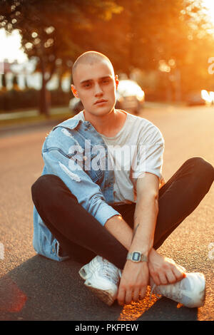 Bel giovane uomo brutale in un elegante giacca denim in grigio di una T-shirt e scarpe bianche si siede su asfalto al tramonto Foto Stock