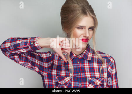 Infelice ragazza triste con rosa Maglietta a scacchi, raccolti updo acconciatura e trucco permanente e guardando la telecamera con faccia insoddisfatta e il pollice verso il basso. Foto Stock