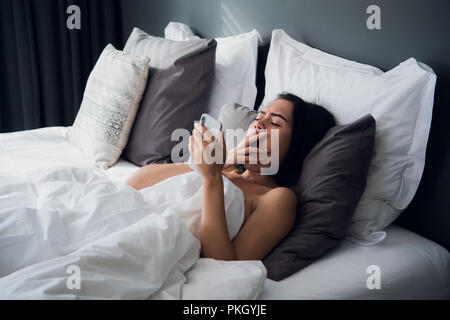 Foto di bella giovane donna in interni di casa utilizzando il telefono cellulare chat. Sbadigliare risiede nel letto. Foto Stock