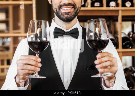 Ritagliato colpo di sorridente sommelier giovane azienda due bicchieri di vino rosso al negozio dei vini Foto Stock