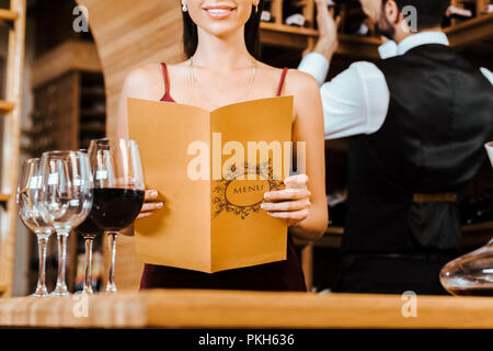 Ritagliato colpo di donna sorridente tenendo premuto menu card presso il negozio dei vini mentre steward tenendo la bottiglia dal ripiano sullo sfondo Foto Stock