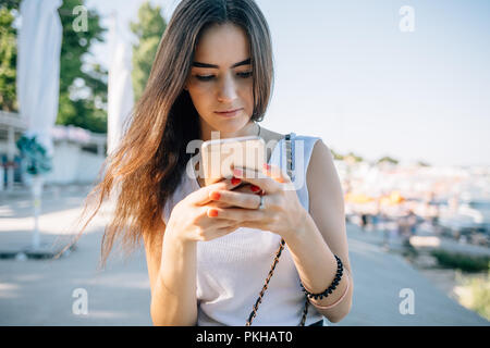 Ritratto di giovane donna con capelli lunghi marrone texting permanente al di fuori sulla strada. Bellissima femmina indossare top bianco utilizzando smart phone al giorno d'estate. Foto Stock