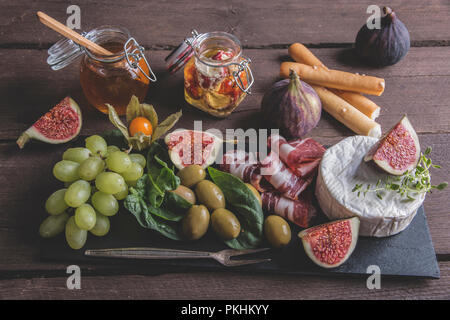 Il prosciutto ,formaggio camembert, fichi,miele, uva, olive on dark serve scheda sopra rustico sfondo di legno Foto Stock