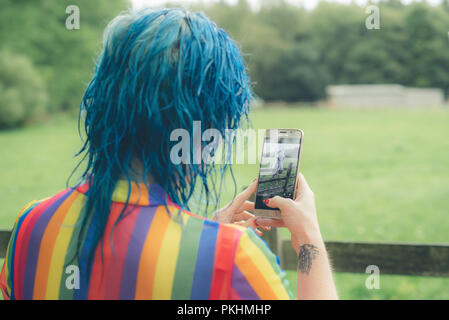 Un blu donna dai capelli, indossando un Gay Pride come top, scatta una foto di una casa sul suo cellulare / Telefono cellulare Foto Stock