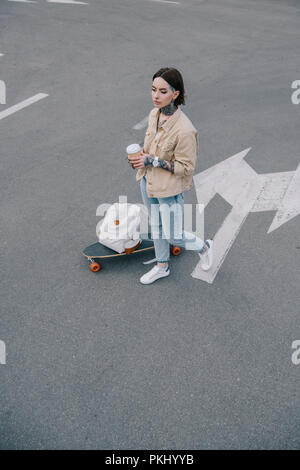 Elevato angolo di visione della donna tatuata con tazza di caffè permanente al parcheggio nei pressi di skateboard e zaino Foto Stock