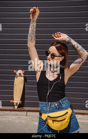 Elegante ragazza tatuata in auricolari ascoltare musica mentre è in piedi con le braccia sollevate Foto Stock