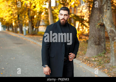 Elegante uomo bello in un mantello nero passeggiate all'aperto su un autunno giornata di sole Foto Stock