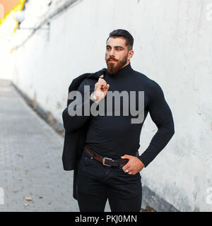 Bel uomo forte con la barba in nero elegante vestiti in posa sulla strada Foto Stock