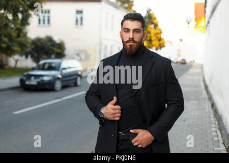 Elegante uomo brutale con la barba in un nero Cappotto alla moda passeggiate sulla strada Foto Stock