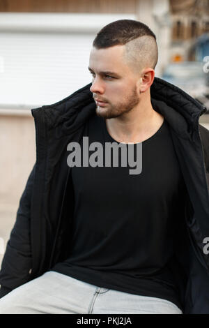 Giovane uomo elegante in una giacca nera si siede sulla strada Foto Stock