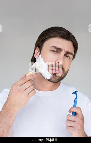 Ritratto di uomo con il pennello e il rasoio in mani mettendo schiuma da barba sulla faccia su sfondo grigio Foto Stock