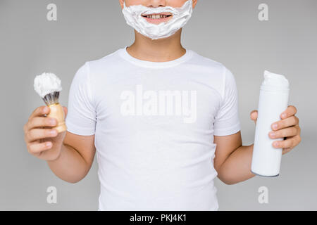 Vista parziale di sorridere preteen boy con pennello, crema e schiuma da barba sulla faccia isolato su grigio Foto Stock