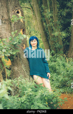 Un blu donna dai capelli, indossando un ponticello blu, si appoggia contro un albero lungo un sentiero forestale Foto Stock