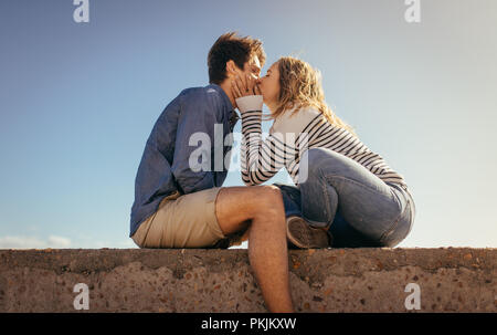 Donna baciare il suo ragazzo seduto su di una parete del mare in una giornata di sole. Coppia in vacanza in atmosfera romantica seduti all'aperto. Foto Stock