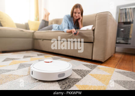 Robotica aspirapolvere pulizia della stanza mentre donna rilassante sul divano. La donna il controllo vuoto con telecomando. Foto Stock