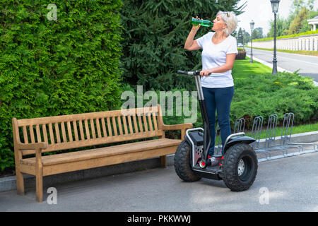 Driver femmina con i capelli bianchi di bere dalla bottiglia verde mentre in piedi su due ruote trasportatore personale accanto al marrone chiaro panca in legno Foto Stock