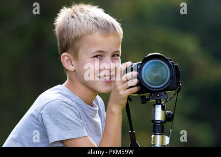 Giovane biondo carino bambino ragazzo di scattare una foto con la fotocamera del treppiede su sfocato copia verde di sfondo spazio. Foto Stock