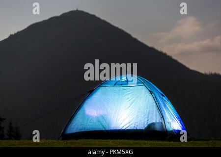 Gli escursionisti turistici tenda illuminata dall'interno sulla verde valle erbosa tra alberi di pino in blu scuro cielo nuvoloso sul distante montagna coperta con fores Foto Stock