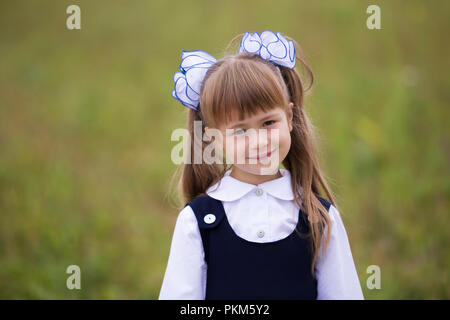 Close-up verticale di graziosi adorabili poco sorridente prima ragazza livellatrice in uniforme scolastica e bianco archi in lunghi capelli biondi sulla luce sfocata copia verde sp Foto Stock