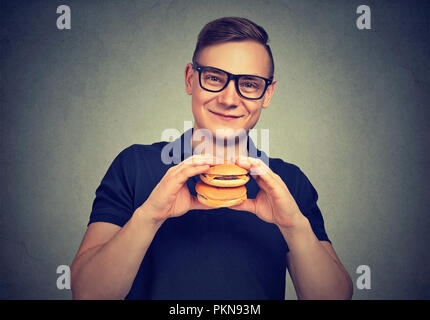 Giovane uomo sorridente in bicchieri holding alcuni hamburger e sorridente in telecamera godendo il fast food Foto Stock