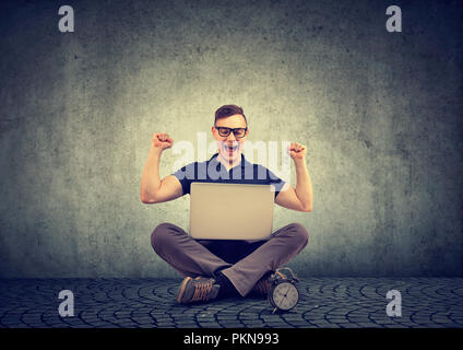 Giovane uomo seduto con laptop e tenendo le mani alzate nella celebrazione di successo essendo eccitato su sfondo grigio Foto Stock