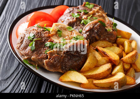 Ossobuco di gambo con una decorazione di fette di patate fritte e pomodori freschi di close-up su una piastra sul tavolo orizzontale. Foto Stock