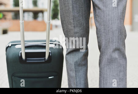 Primo piano di un giovane imprenditore caucasico in un elegante abito grigio tirando una valigia trolley per la maniglia sulla strada Foto Stock