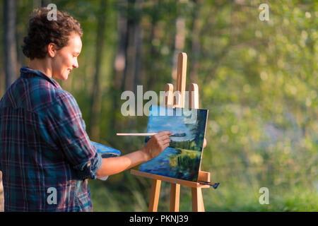 Giovane donna graziosa artista disegna dipinge un quadro di un lago su open plain air all'aperto Foto Stock