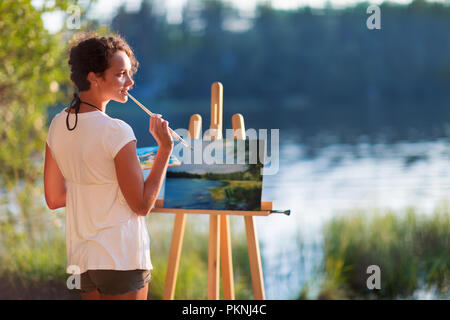 Giovane donna graziosa artista disegna dipinge un quadro di un lago su open plain air all'aperto Foto Stock