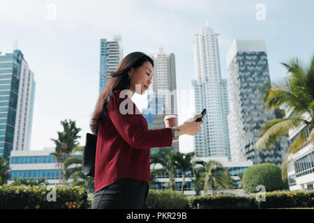 Felice femmina cinese manager messaging sul telefono cellulare, a bere caffè. Successo imprenditrice asiatica è raggiungibile a piedi in ufficio al mattino. Foto Stock