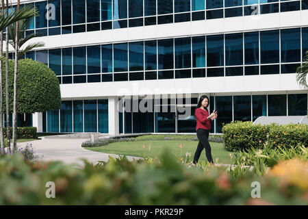 Felice femmina cinese manager messaging sul telefono cellulare, a bere caffè. Successo imprenditrice asiatica è raggiungibile a piedi in ufficio al mattino. Foto Stock
