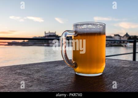 Vetro freddo boccale di birra con schiuma sullo sfondo il castello di Bratislava e il fiume Danubio Foto Stock