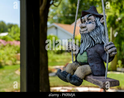 Rustico maschio cercando Gnome con la barba bianca e un cappello su uno swing con giardini in background Foto Stock