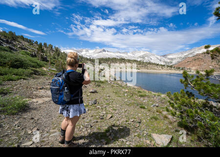 Femmina bionda backpacker escursionista si ferma per scattare una foto di un lago alpino in montagna. 20 bacino dei Laghi escursione nelle montagne della Sierra Nevada di Cali Foto Stock