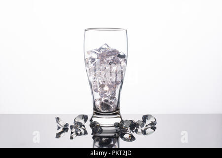 In vetro trasparente riempita con diamanti su uno sfondo bianco con una riflessione al di sotto di esso. Diamanti allentati al di sotto di esso. Foto Stock