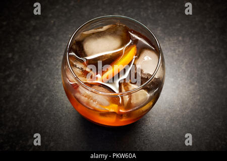 Un isolato di vetro luminoso giallo / marrone bevanda alcolica con ghiaccio e fettine di arancia contro uno sfondo nero Foto Stock