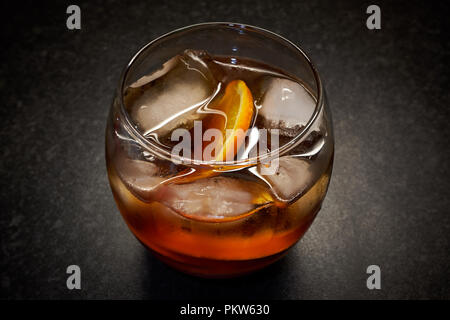 Un isolato di vetro luminoso giallo / marrone bevanda alcolica con ghiaccio e fettine di arancia contro uno sfondo nero Foto Stock