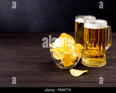 2 tazza di vetro della luce della birra e patatine fritte in un recipiente di vetro al buio su un tavolo di legno. Foto Stock