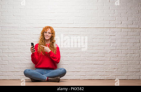 Giovani redhead donna seduta su un muro di mattoni a parlare al telefono molto felice puntando con mani e dita Foto Stock
