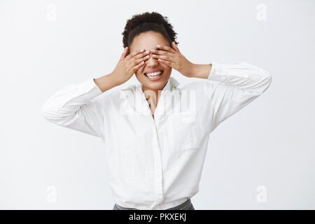 Tiro al coperto di affascinanti carefree afro-americano datore di lavoro femminile con la camicia bianca che copre gli occhi con le palme, essendo accecato durante la riproduzione di peek-a-boo e sorridente ampio alla fotocamera sulla parete grigia Foto Stock