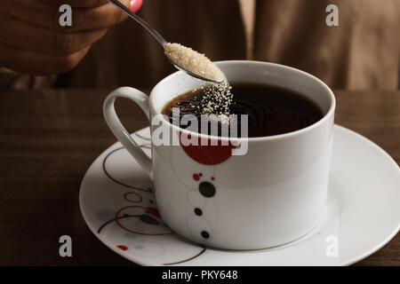 Close-up di una donna di mettere un cucchiaio di zucchero di canna nella sua tazza di caffè. Foto Stock