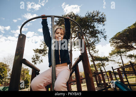 Ritratto di giovane ragazza appeso su un telaio di arrampicata mediante le sue braccia. Bambina giocare nel parco giochi. Foto Stock