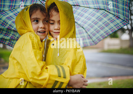 Adorabili ragazze che indossano tute impermeabili che abbraccia ogni altra sotto l'ombrello. Gemelle godendo all'aperto sul giorno di pioggia. Foto Stock