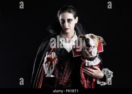 La donna e il suo cane in simili vampire costumi per Halloween. Giovani femmine con un bicchiere di rosso e di bere il suo cucciolo vestito nella stessa costume di Dracula Foto Stock