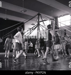 Degli anni Cinquanta, storiche, i giovani della scuola bambini saltando o ballando intorno un maypole all'interno di una sala durante una classe PE, Inghilterra, Regno Unito. Foto Stock