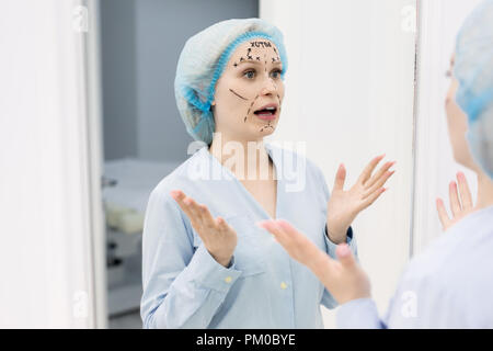 Confuso femmina in camice bianco in piedi di fronte ad uno specchio e guardando il suo viso diviso in zone con un evidenziatore nero Foto Stock