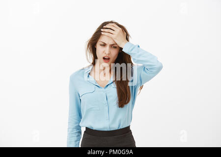 Vita-up shot di orinato cupo donna europea in blu camicetta, azienda palm sul fronte e accigliata, guardando verso il basso, con la sensazione di disordine, di mal di testa o che soffrono di postumi della sbornia oltre il muro grigio Foto Stock